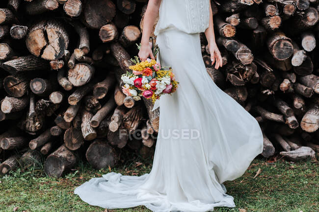 Dame méconnaissable en robe blanche et avec un bouquet nuptial tournant tout en dansant près des piles de bûches pendant le mariage à la campagne — Photo de stock