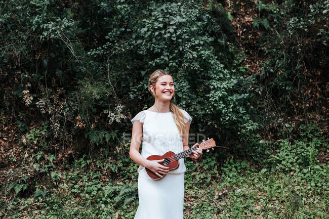 Счастливая молодая женщина в белом платье улыбается и смотрит в сторону, стоя возле зеленых кустарников и играя на укулеле во время свадьбы в сельской местности — стоковое фото