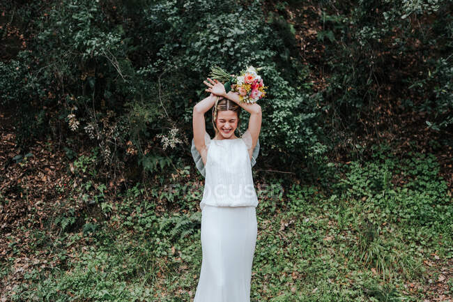 Deliziosa signorina in abito bianco sorridente ad occhi chiusi e mani incrociate con bouquet da sposa sopra la testa mentre in piedi vicino a cespugli verdi in campagna — Foto stock
