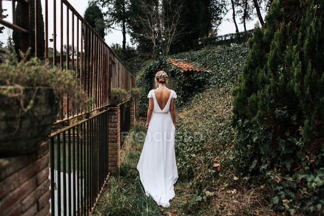 Обратный вид на неузнаваемую женщину в элегантном белом платье, идущую возле металлического забора в день свадьбы в зеленом саду — стоковое фото