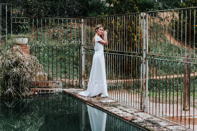 Вид сбоку на молодую женщину в белом свадебном платье, стоящую на выветренной границе возле металлического забора и спокойный бассейн в саду — стоковое фото