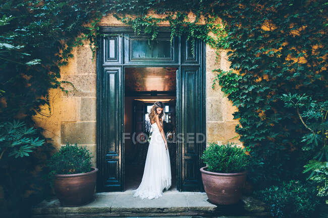 Back view of young sensual bride in white wedding dress standing in doorway of old stone building covered with green ivy and potted plants — Stock Photo