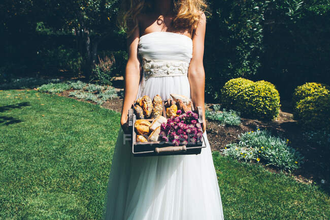 Невпізнавана молода наречена в елегантній білій весільній сукні, що стоїть на зеленому газоні в саду і тримає кошик зі свіжими булочками і хлібом — стокове фото