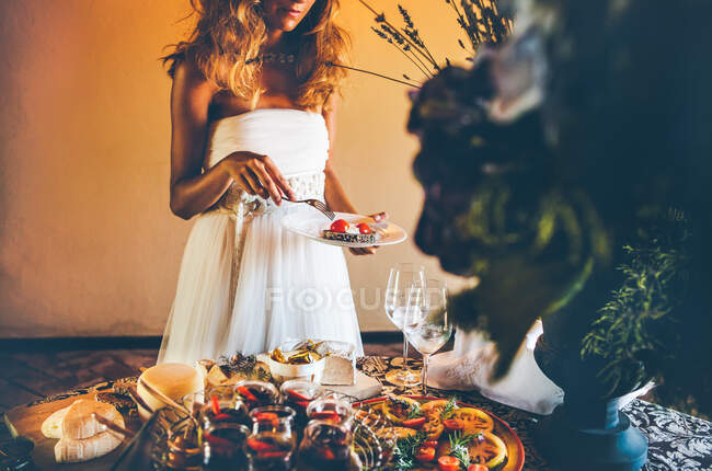 Anonymous young bride in elegant white wedding dress holding plate and fork while standing near banquet table with wineglasses and tray with cheese and bread and fruit desserts — Stock Photo