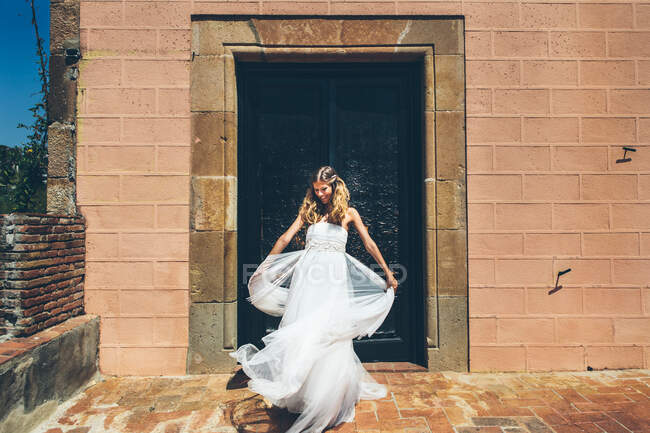 Cheerful elegant young bride in stylish white wedding dress spinning around near entrance of old stone building during wedding celebration — Stock Photo
