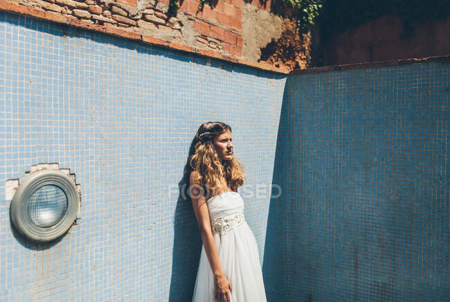 Vista lateral da sensual jovem noiva loira em elegante vestido de noiva branco de pé sozinho na piscina rasca vazia e olhando para longe — Fotografia de Stock