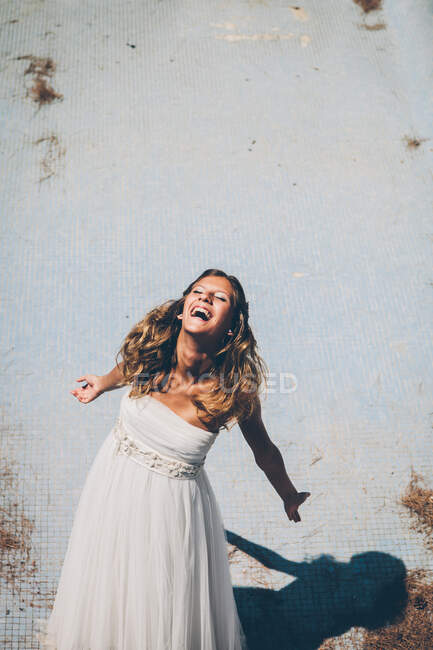 From above view of happy young blond bride in elegant white wedding dress standing alone on shabby bottom of pool with hands apart and laughing loudly with closed eyes — Stock Photo