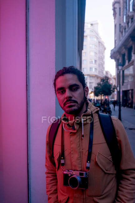 Handsome ethnic male photographer in casual clothes with black backpack and bun of hair standing on street near shop window with neon glow and looking at camera with film camera hanging on neck — Stock Photo