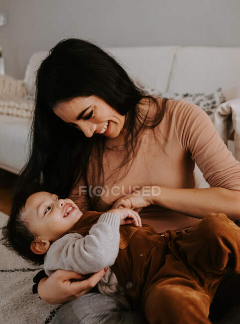 From above view of cheerful young mother in casual clothes hugging cute smiling little son while sitting with crossed legs on carpet on floor near decorated Christmas tree — Stock Photo