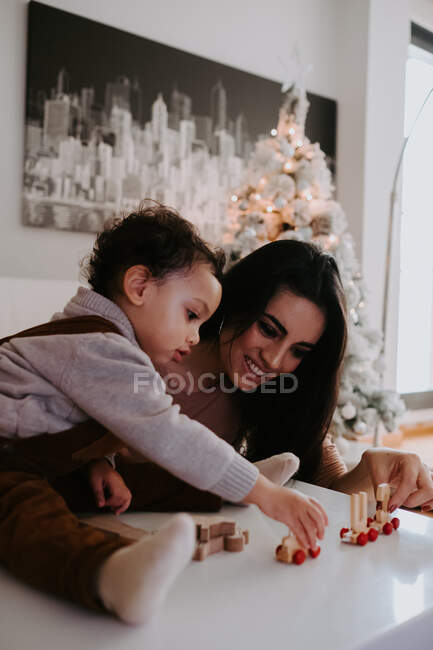 Madre sonriente sentada cerca de la mesa en la sala de estar decorada y jugando con un lindo hijo pequeño con juguetes de madera - foto de stock