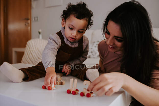 Lächelnde Mutter sitzt am Tisch im dekorierten Wohnzimmer und spielt mit niedlichem kleinen Sohn mit Holzspielzeug — Stockfoto