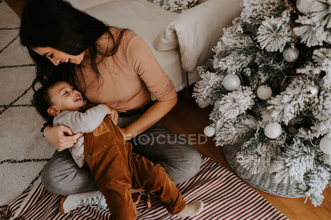 Сверху вид веселой молодой матери в повседневной одежде обнимающей милого улыбающегося маленького сына, сидящего со скрещенными ногами на ковре на полу возле украшенной елки — стоковое фото