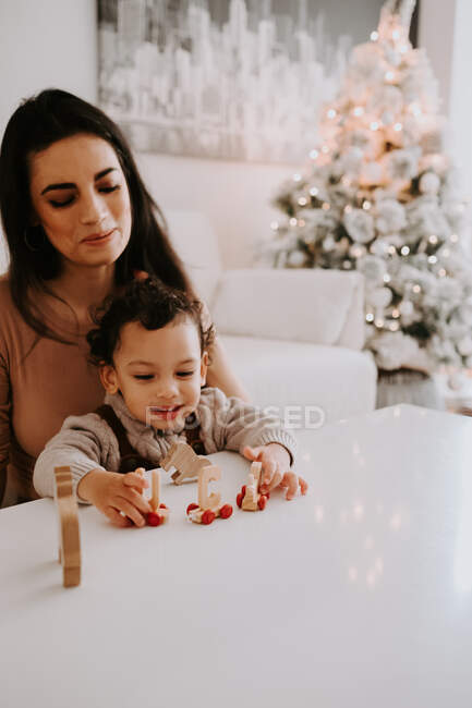 Sorridente madre seduta vicino al tavolo in salotto decorato e giocare con il piccolo figlio carino con giocattoli in legno — Foto stock