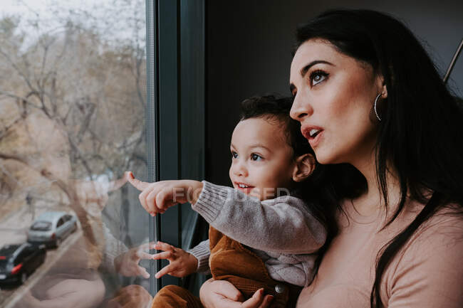 Вид збоку люблячої матері в повсякденному одязі тримає милий маленький син на руках, стоячи біля вікна вдома і вказуючи геть — стокове фото