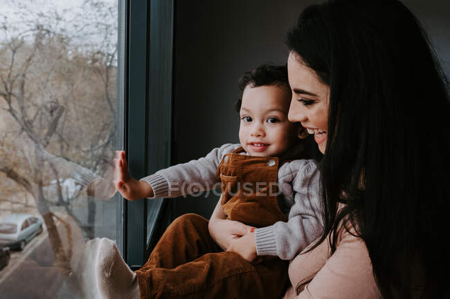 Vista lateral de la madre cariñosa en ropa casual sosteniendo lindo hijo pequeño en las manos mientras está de pie cerca de la ventana en casa y apuntando hacia otro lado - foto de stock
