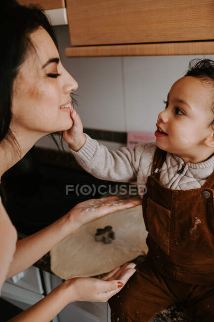 Side view of cheerful little boy in casual clothes with flour on cheeks helping mother cooking pastry in modern kitchen — Stock Photo