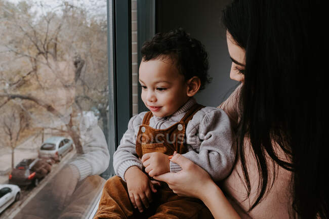 Vista lateral de la madre cariñosa en ropa casual sosteniendo lindo hijo pequeño en las manos mientras está de pie cerca de la ventana en casa y apuntando hacia otro lado - foto de stock