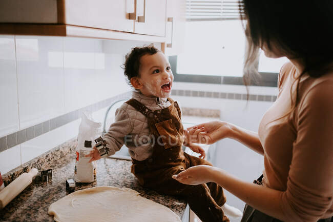 Side view of cheerful little boy in casual clothes with flour on cheeks helping mother cooking pastry in modern kitchen — Stock Photo