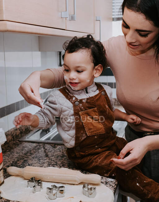 Боковой вид веселой молодой матери в повседневной одежде, готовящей тесто со скалкой с сыном, стоящим на стуле на светлой кухне — стоковое фото