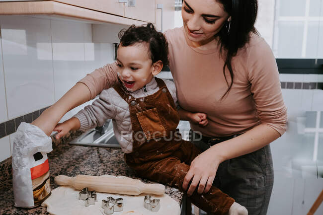 Side view of cheerful young mother in casual clothes preparing dough with rolling pin with son standing on chair in light kitchen — Stock Photo