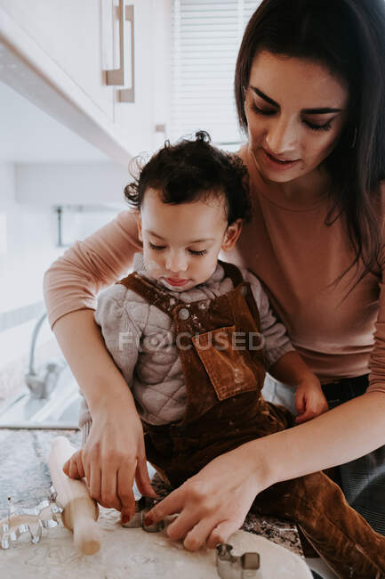 Vista lateral de la alegre madre joven en ropa casual preparando la masa con rodillo con el hijo de pie en la silla en la cocina ligera - foto de stock