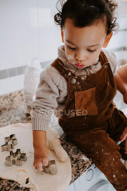 Little cute boy with curly hair in casual clothes smeared with flour cooking cookies with metal baking molds at table in kitchen — Stock Photo