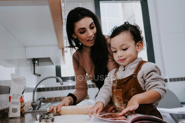 Vista lateral de la alegre madre joven en ropa casual preparando la masa con rodillo con el hijo de pie en la silla en la cocina ligera - foto de stock