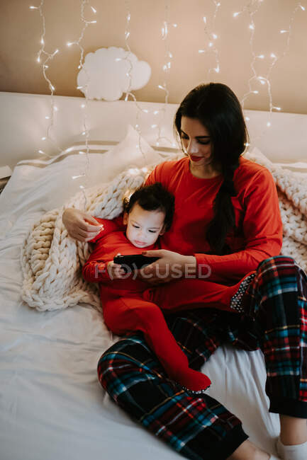 Зверху від позитивної матері в теплому сплячому одязі, що обіймає чарівного маленького сина в червоній теплій піжамі, дивлячись мультфільми на смартфон у світлій декорованій спальні — стокове фото