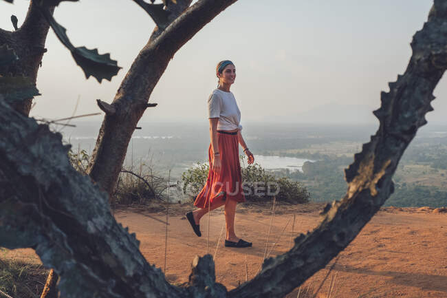 Vista lateral de la mujer en camiseta casual y falda caminando mirando hacia otro lado en una colina arenosa mientras disfruta de la puesta del sol y viaja por Sri Lanka - foto de stock