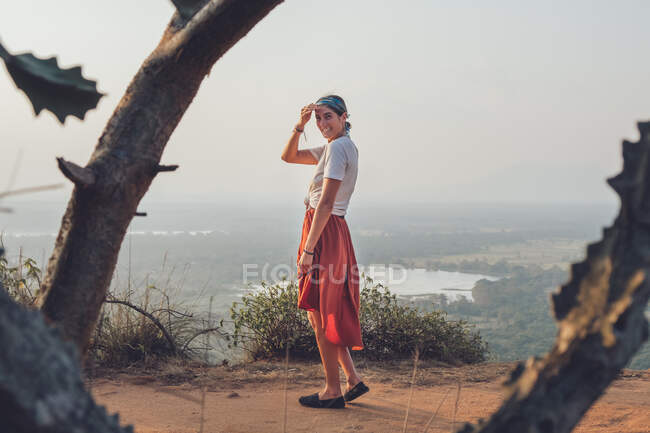 Vista lateral de la mujer en camiseta casual y falda de pie sobre una colina arenosa y cubriendo la cara mientras disfruta de la puesta del sol y viaja por Sri Lanka - foto de stock