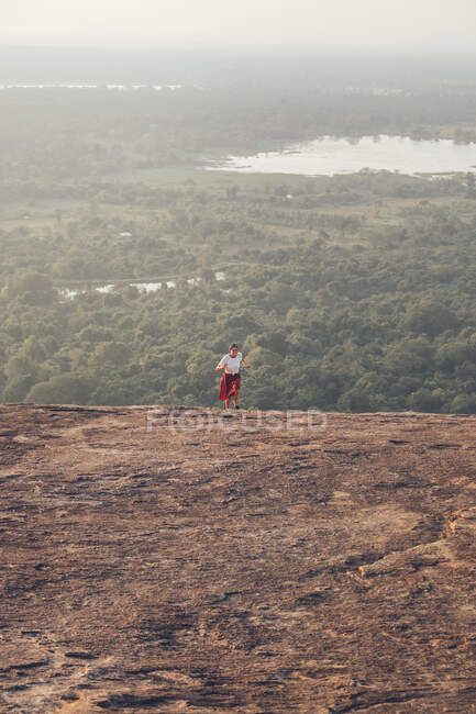 Desde arriba de viajar hembra en ropa casual corriendo cuesta arriba en el fondo del valle con bosque y lago durante las vacaciones de verano en Sigiriya - foto de stock