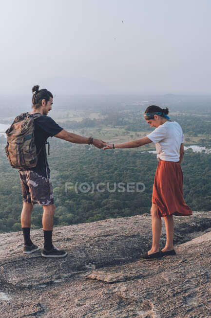 Desde arriba vista lateral de un par de hipsters en ropa casual tomados de la mano y disfrutando de la puesta de sol en el borde de piedra de la colina mientras viaja alrededor de Sigiriya en Sri Lanka - foto de stock