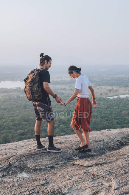 From above side view of couple of hipsters in casual wear holding hands and enjoying sunset on stone edge of hill while traveling around Sigiriya in Sri Lanka — Stock Photo