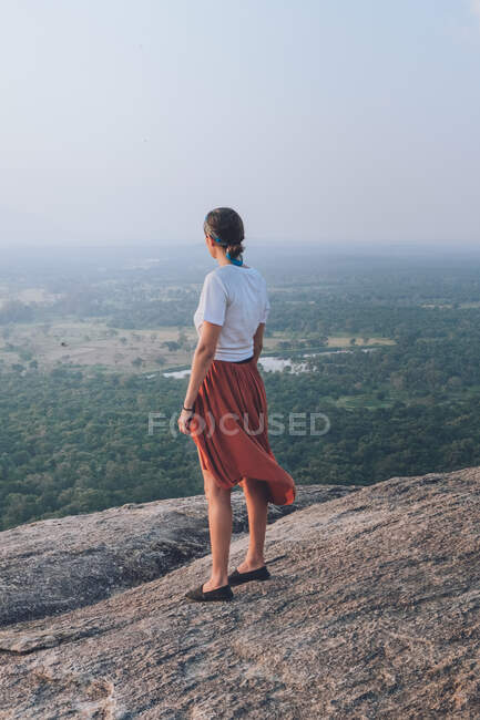 Vista laterale di donna viaggiante irriconoscibile in abiti casual in piedi in una collina rocciosa sullo sfondo della valle con foresta e lago durante le vacanze estive a Sigiriya — Foto stock
