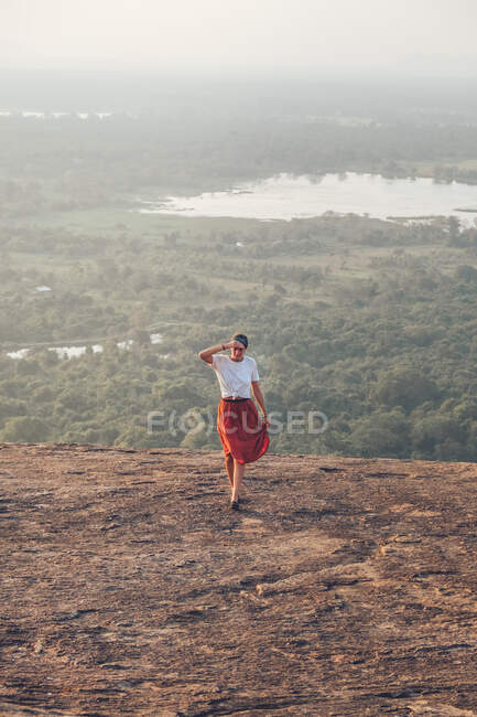 Сверху путешествующая женщина в повседневной одежде, идущая в гору на фоне долины с лесом и озером во время летнего отдыха в Сигирии — стоковое фото