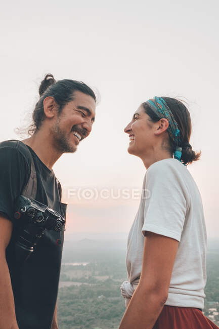 Low angle side view of laughing couple of travelers in casual wear standing on background of green valley landscape and looking at each other — Stock Photo