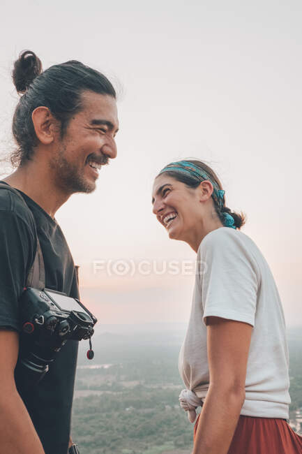 Low angle side view of laughing couple of travelers in casual wear standing on background of green valley landscape and looking at each other — Stock Photo