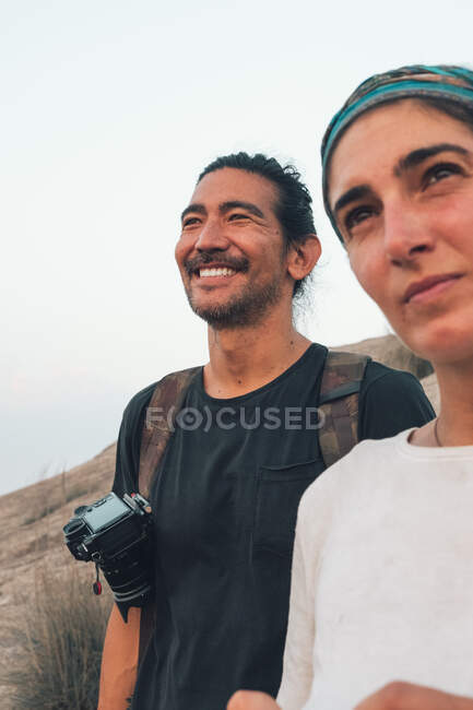 Low angle of happy couple of travelers in casual wear standing on background of green valley landscape and looking away — Stock Photo