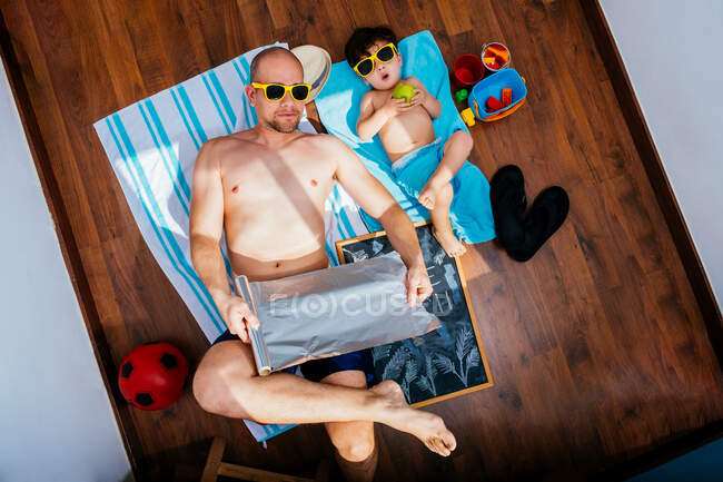 Desde arriba de padre e hijo positivo en gafas de sol amarillas y bañadores tumbados en el suelo sobre toallas y soñando con vacaciones de verano en la playa durante la cuarentena - foto de stock