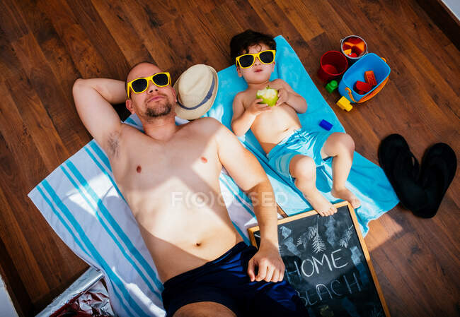 From above view of cheerful boy in yellow sunglasses and blue swimwear smiling while feeding father apple lying on towels together having beach at home — Stock Photo
