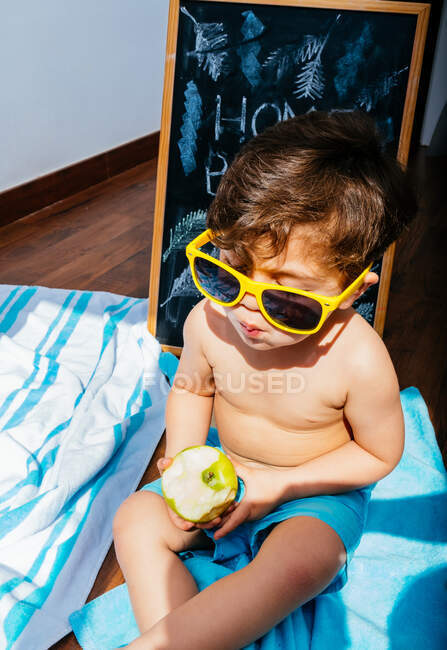 De cima vista de menino alegre em óculos de sol amarelos e azul swimwear comer maçã enquanto deitado em toalha no chão tendo praia em casa em quarentena — Fotografia de Stock