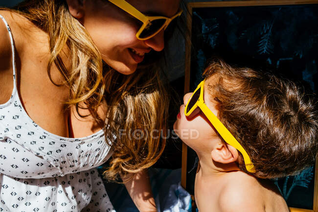 Vista lateral de madre positiva en vestido blanco y gafas de sol amarillas tocando narices con hijo en las mismas gafas de sol en luz solar brillante - foto de stock