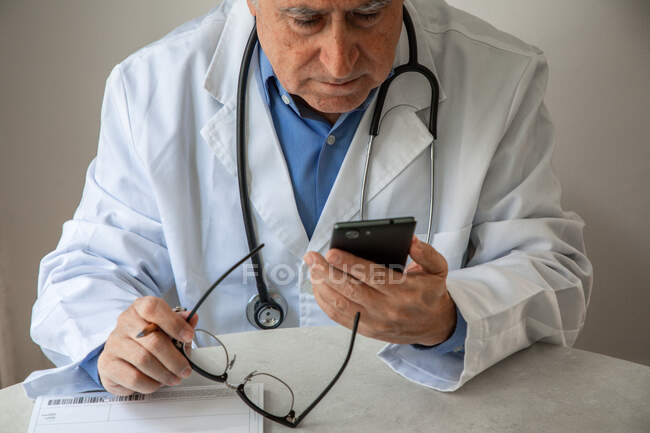 Älterer Arzt im Arztkittel sitzt am Tisch und schaut aufs Smartphone — Stockfoto
