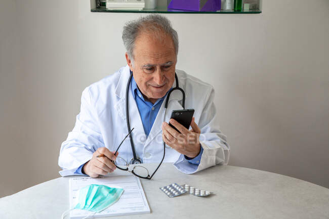 Médecin homme âgé en robe médicale assis à table et regardant smartphone — Photo de stock
