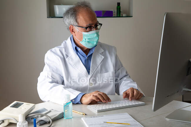 Старший лікар у масці для обличчя та медичній сукні сидить за столом та друкує на клавіатурі — стокове фото