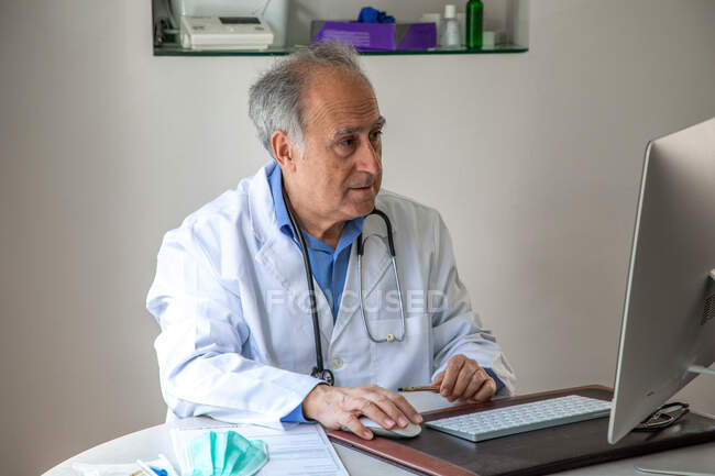 Лікар - старший чоловік у медичній сукні сидить за столом і дивиться на екран комп 
