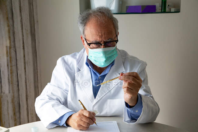 Мужчина-врач в защитной маске пишет на бумаге и смотрит на термометр — стоковое фото
