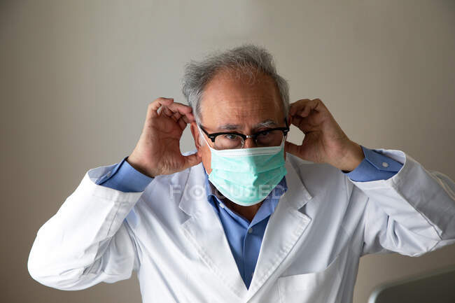 Médico idoso em vestido médico branco colocando máscara facial em — Fotografia de Stock