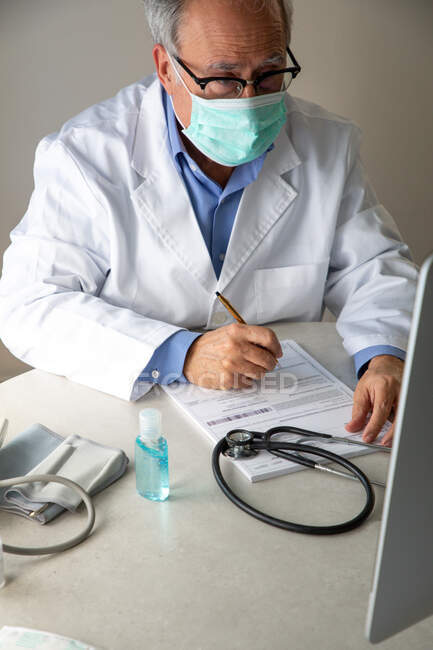 Médico general masculino con máscara protectora y bata médica sentado en la mesa e informe de escritura - foto de stock