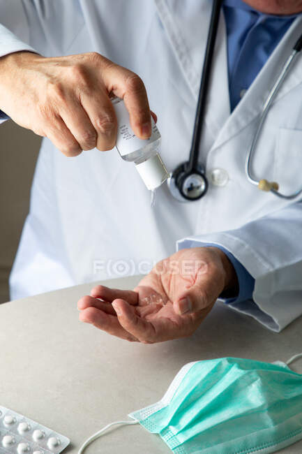 Male therapist in medical gown sitting at table in hospital and sanitizing hands with antiseptic — Stock Photo